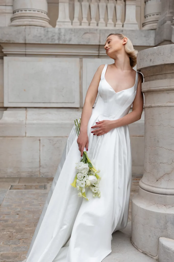 Frau im weißen Kleid lehnt an einer Säule und hält einen Blumenstrauß