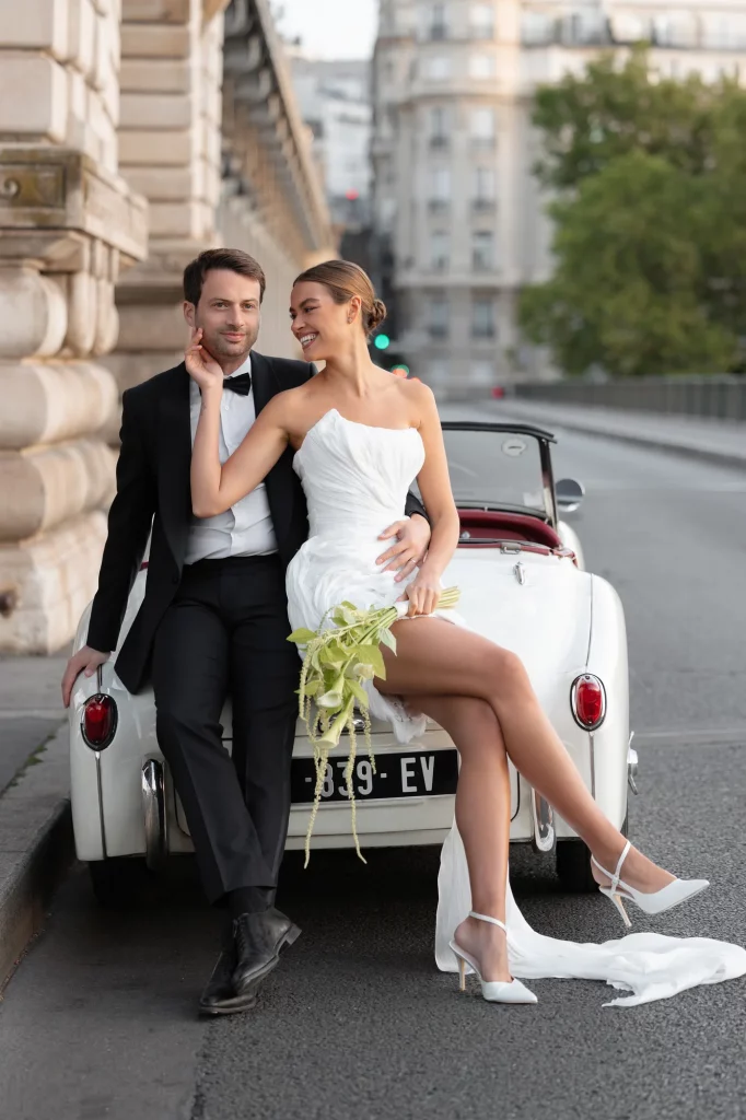 Zwei Personen sitzen auf einem weißen Auto mit Blumenstrauß und Stadtstraße im Hintergrund